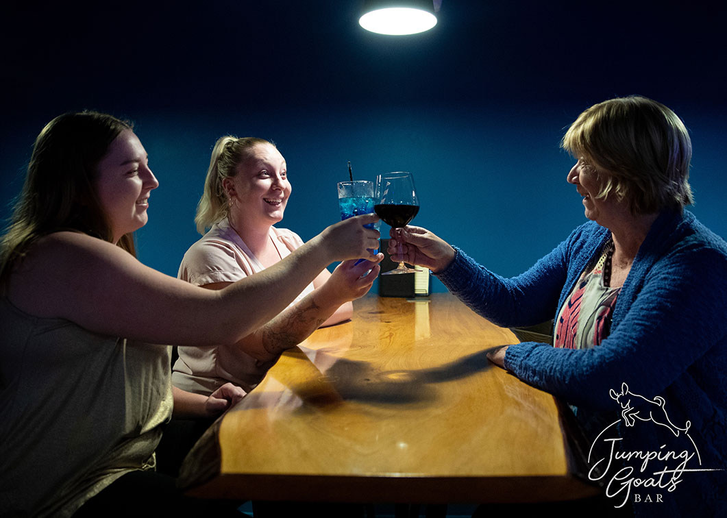 Three friends toasting with wine and cocktails over a timber slab table